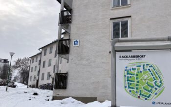 Bilden föreställer studentbostadsområdet Rackarberget i Uppsala. Till höger en områdeskarta över området. Bakom den en smutsvit husfasad med en Uppsalahem-skylt på. I bakgrunden syns flera andra hus i samma färg. Marken är täckt av snö.