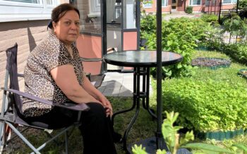 Kvinna på en grönskande uteplats. Hon sitter på en stol till vänster i bilden och ser ledsen ut. Hon har händerna i knät, har en leopardmönstrad topp, svarta byxor och mörkt hår uppsatt i en knut.