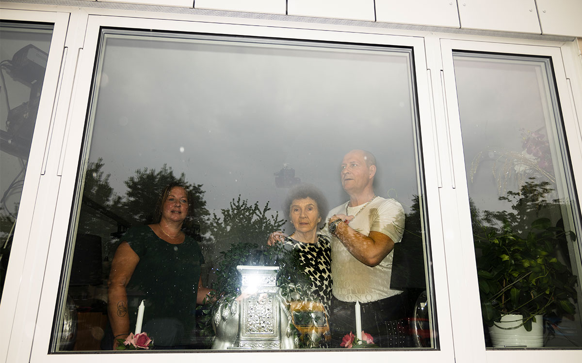 En ung kvinna, en man och en äldre kvinna står i ett fönster utan persienner.