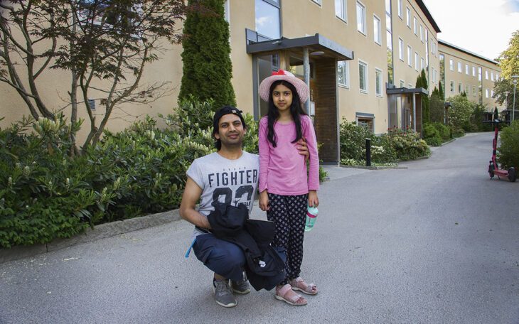 Yasir Shah sitter på huk bredvid dottern Zeena Iman med mobilen, som står upp. Han har på sig en bakåtvänd svart keps och en ljusgrå tshirt med tryck. Hon har långt mörkt hår och har på sig en ljusrosa solhatt med en rosa dekoration, en rosa långärmad tröja och mörka byxor med ljust mönster. De står framför en länga hyreshus i tre våningar med sandfärgad putsad fasad. Yasir Shah hade inte koll på att avvecklingen av 3G-nätet och 2G-nätet påverkar porttelefoner.