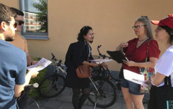 Åse Richard i röd kortärmad topp och jeansshorts, långt grått hår och glasögon, berättar ooch gestikulerar omgiven av deltagarna i stadsvandringen. De står intill en aprikosfärgad husvägg och i ett fönster speglas laxrosa bostadshus och grönska.