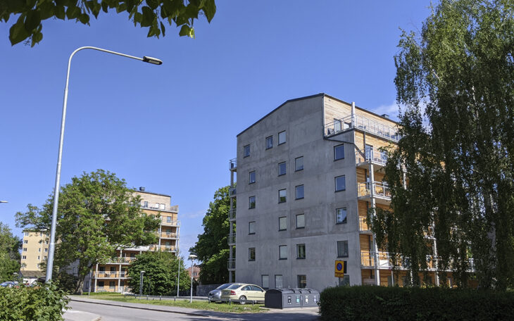 Två hus i sex-sju våningar med avskalade gråbeige betongfasader mot gatan och träfasad med balkonger in mot gårdarna. Ytterligare ett hus i bakgrunden. Gränskande träd, bland annat en stor björk längst till höger i bild. Det är ännu oklart om investeringstödet för husen i Rickomberga kommer att betalas ut.