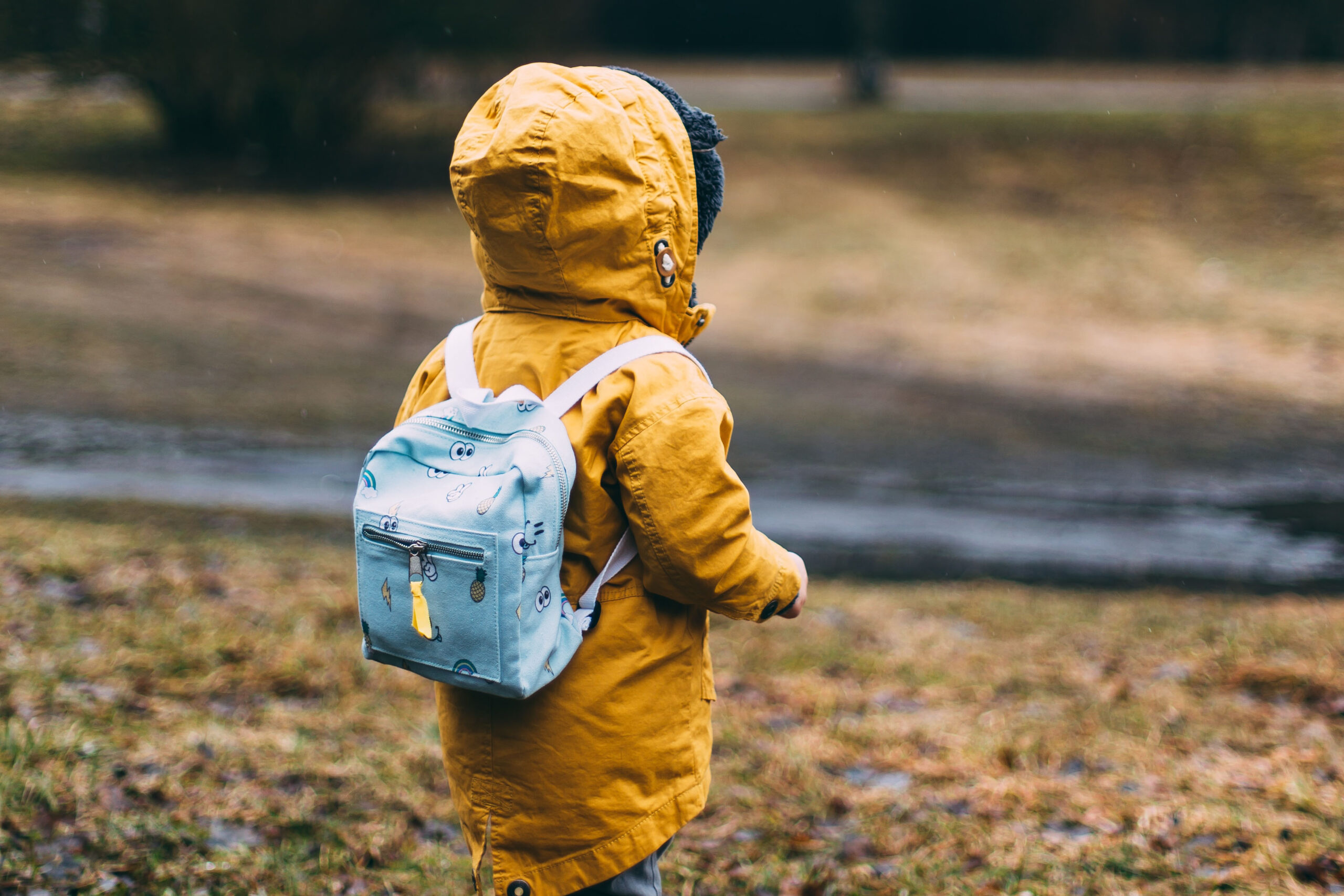 Ett litet barn iklädd gul jacka och med en ljusblå ryggsäck på ryggen. Bilden är tagen så att man inte ser barnets ansikte.