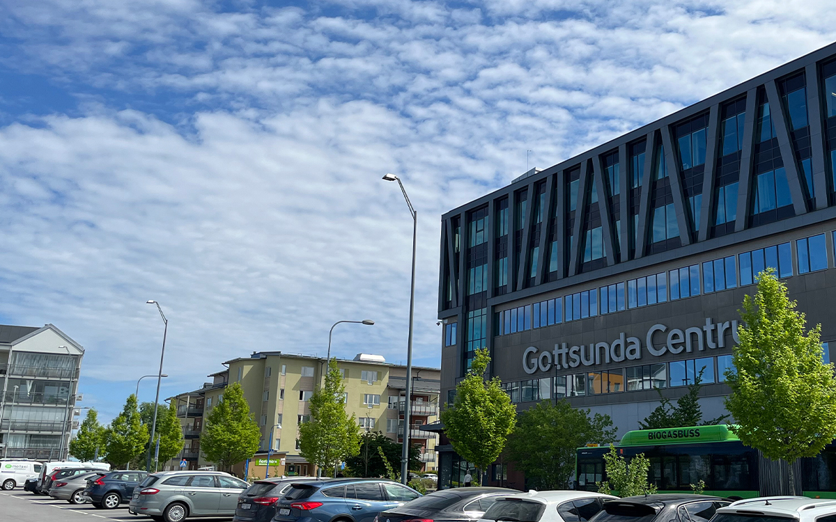 En centrumbyggnaderna med bokstäverna Gottsunda C syns till höger. Framför byggnaden är bilar på en parkeringsplats. I bakgrunden några hus. Himlen är blå med vita moln. Några gröna träd framför centrumbyggnaden.