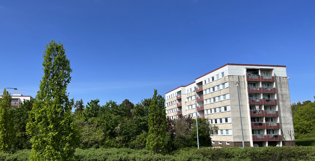 Till höger en huskropp med sex våningar. Till vänster om den gröna träd och buskar. Klarblå himmel.