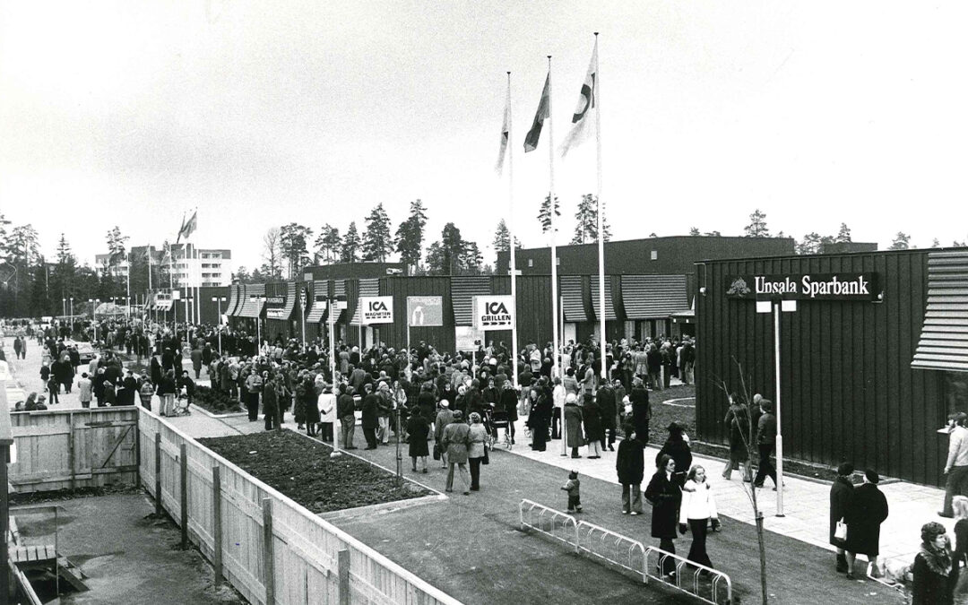En svartvit bild på ett köpcentrum på 1970-talet, med en massa folk utanför.