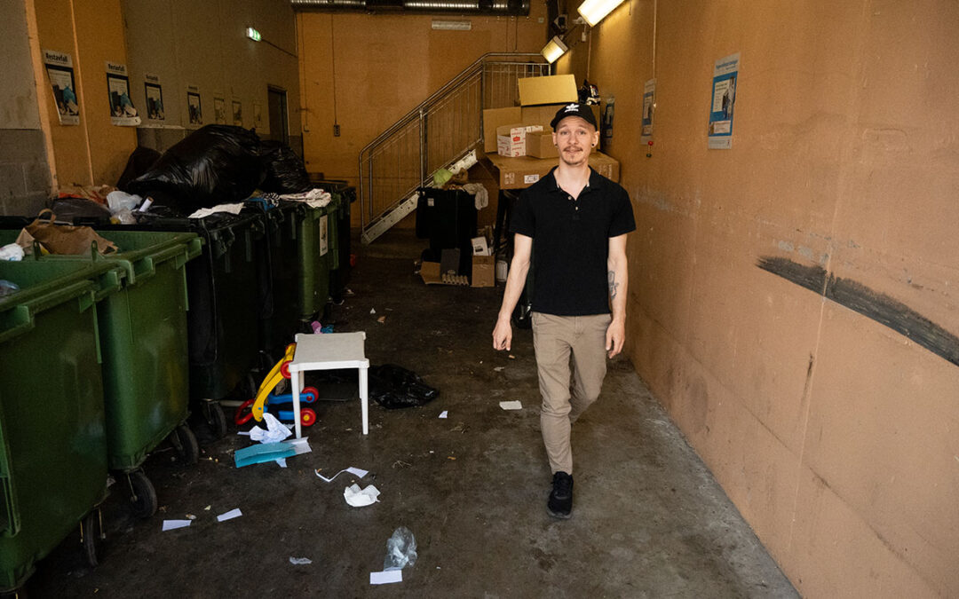 En ung man som går i ett överfullt soprum med diverse avfall både i överfulla soptunnor och på golvet.