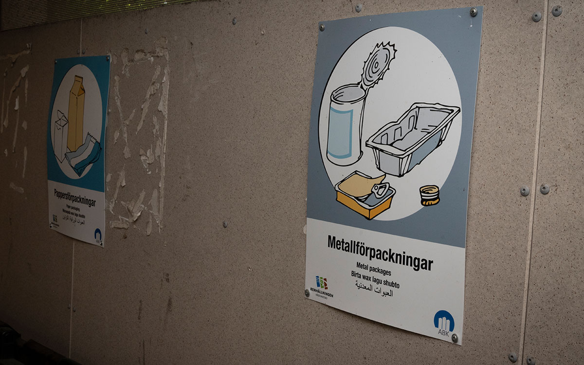 Två skyltar på en vägg som visar vilken typ av avfall som ska läggas i vilken behållare: "Metallförpackningar/Metal packages/Birta wax lagu shubto" och motsvarande skylt för pappersförpackningar, även denna med text på svenska, engelska, somaliska och arabiska.