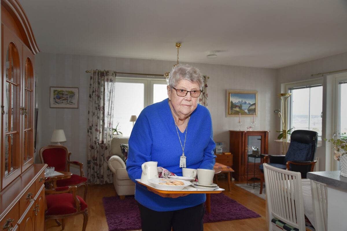 En äldre dam i blå tröja som bär en bricka med kakor och bullar.