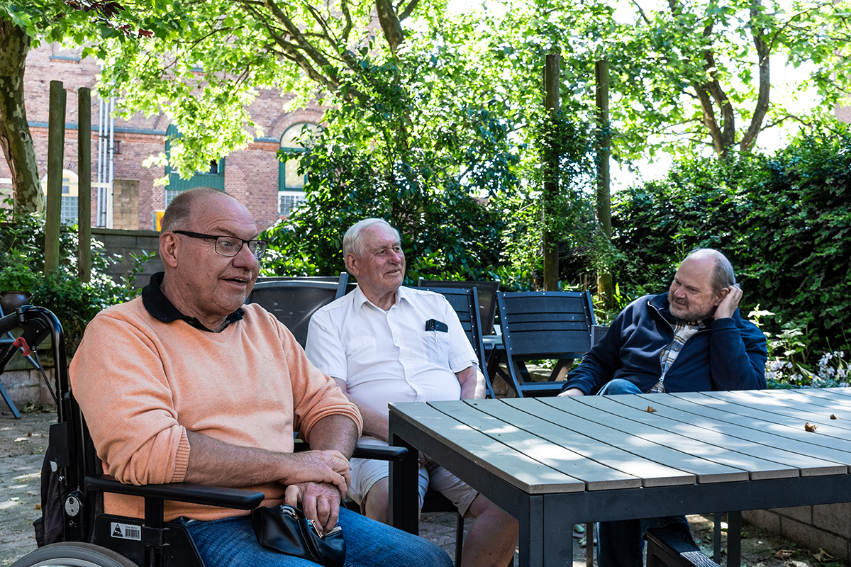 Tre äldre män sitter i skuggan utomhus.