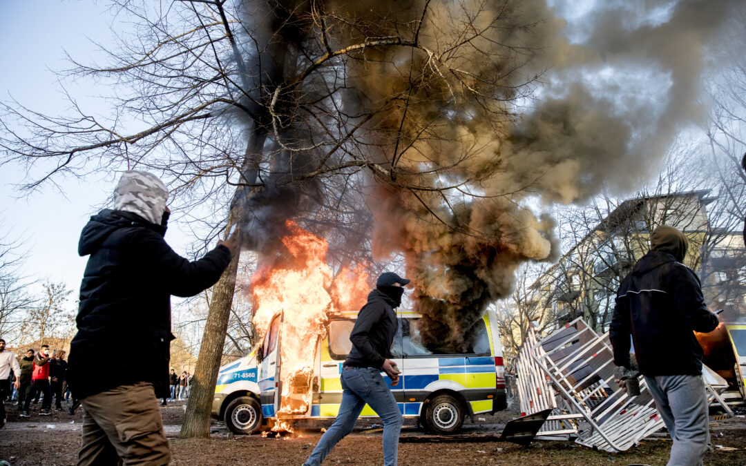 En polisbil i brand och tre maskerade personer som fotar och bär på stenar.