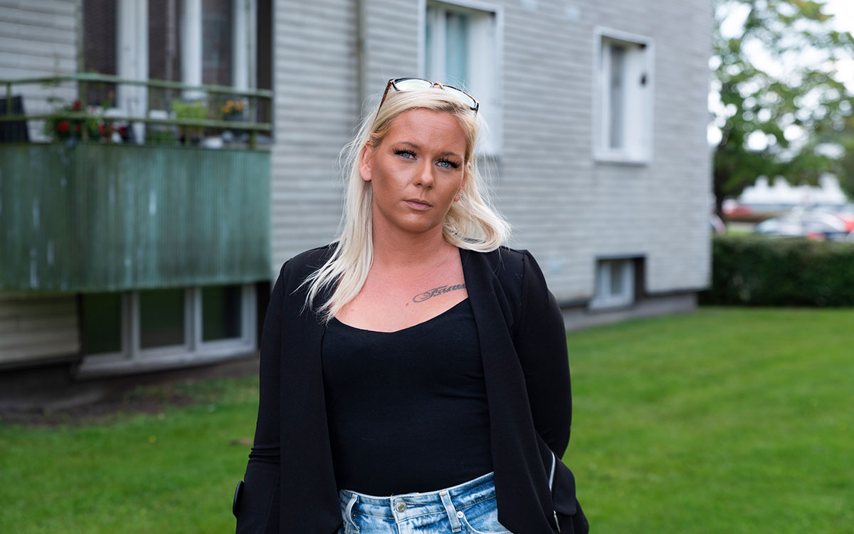 En kvinna med blont hår och blå ögon iförd jeansshorts och svart linne som står framför ett bostadshus.