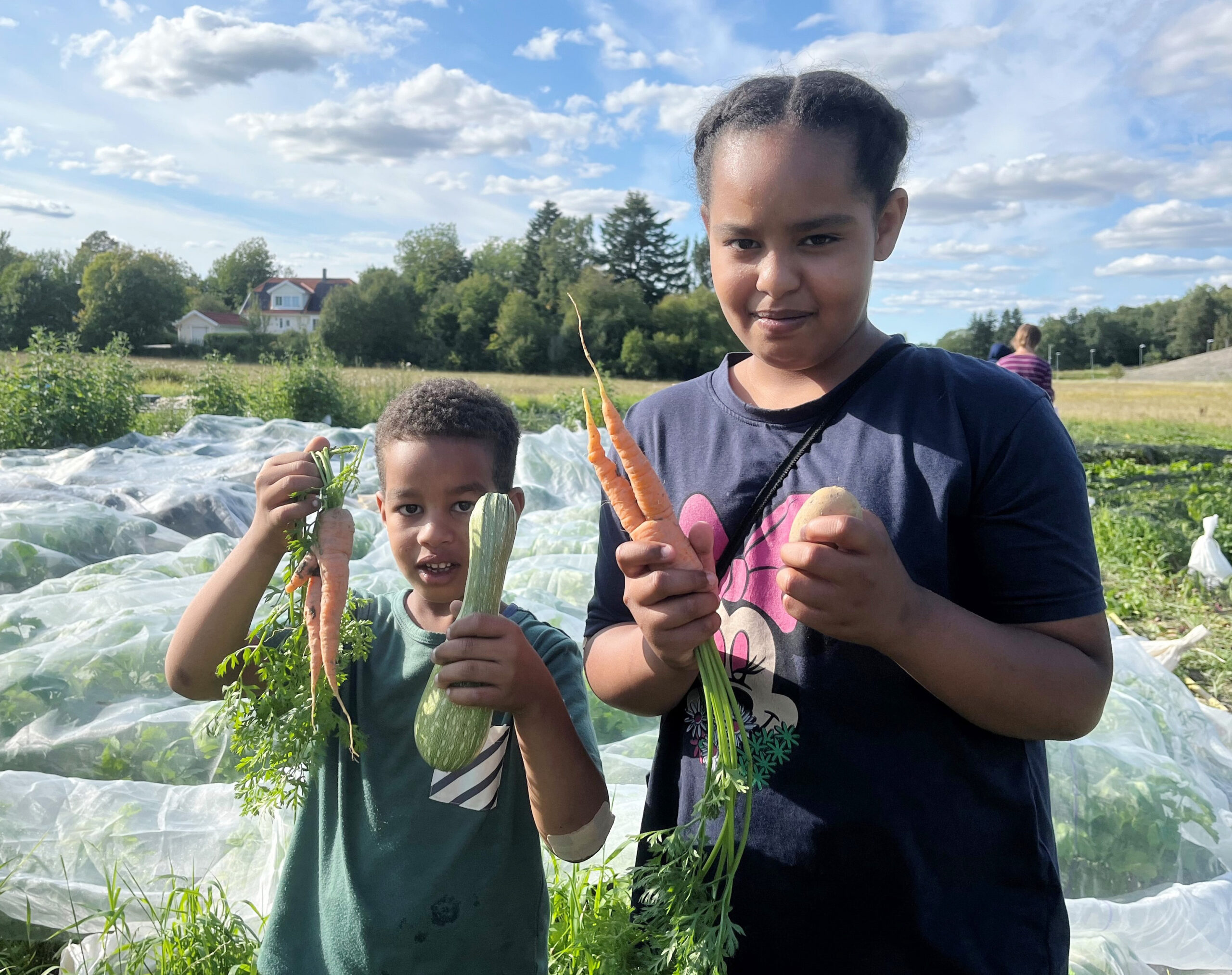 Två barn, En mindre mörkhyad pojke står till vänster i ett odlingland. Framför sig håller han jordiga morötter och en zucchini. Till höger om honom i bilden står hans 12-åriga syster, som håller morötter med ena handen och potatis i den andra. Båda tittar in i kameran.