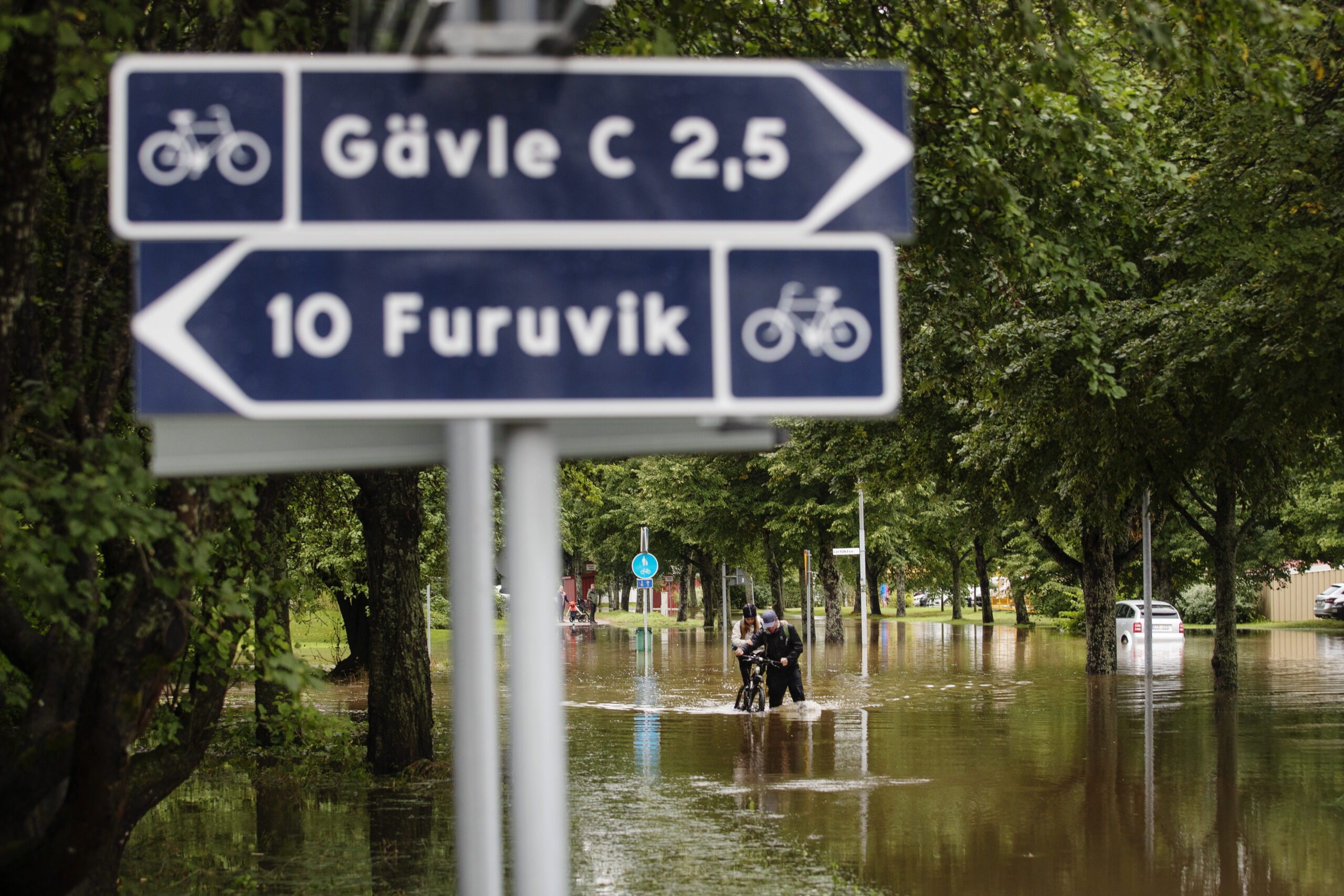 Två cykelvägsskyltar i förgrunden. På den ena står det Gävle 2,5 km, och på den andra Furuvik 10 km. I bakgrunden en översvämmad gata.