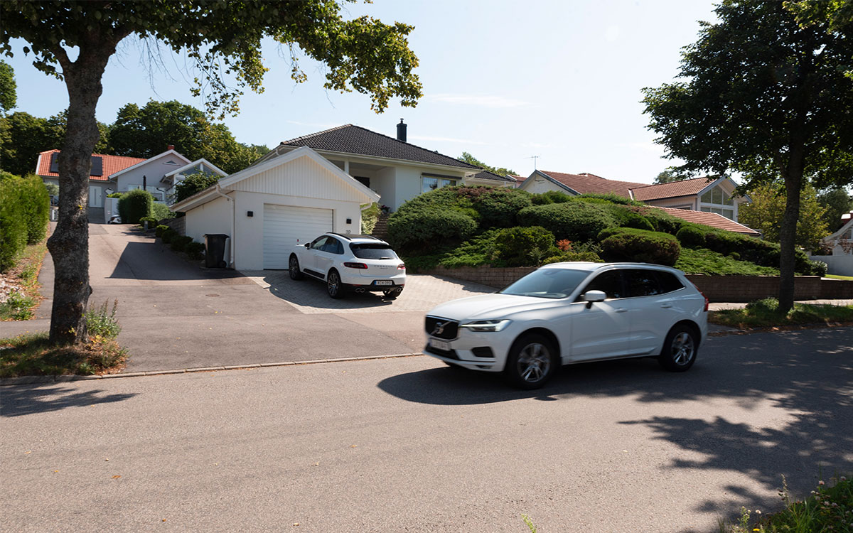 En villagata med en vit Volvo-bil i rörelse och en Porsche parkerad på en garageuppfart.