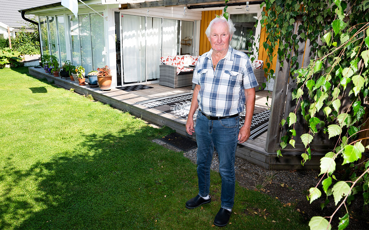 En äldre man i jeans och rutig skjorta som står vid en uteplats vid en villa med gul träpanel och en svensk flagga på fasaden.