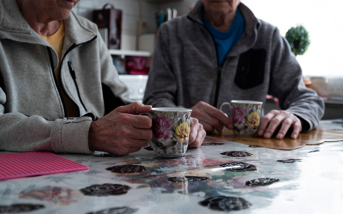 Två äldre personer som sitter vid ett köksbord och håller i varsin blommig kaffemugg som står på ett bord med blommig duk.