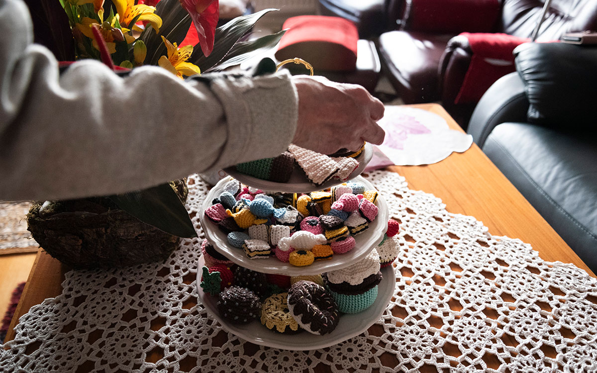 Ett kakfat i tre våningar som står på ett bord i ett vardagsrum. Fatet innehåller virkade föremål som ser ut som engelsk konfekt, munkar, chokladbollar och andra bakverk. En hand som tillhör en äldre person tar eller lägger något på fatet.