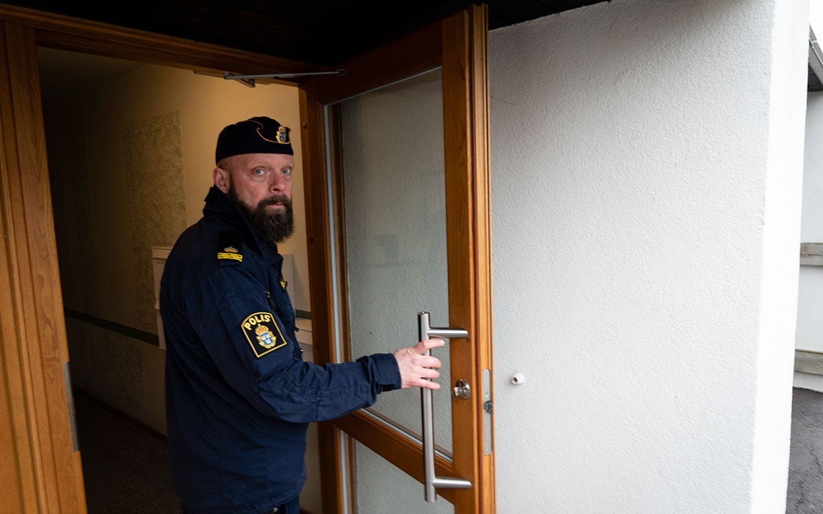 En man med skägg och polisuniform med båtmössa som öppnar en port till ett lägenhetshus.