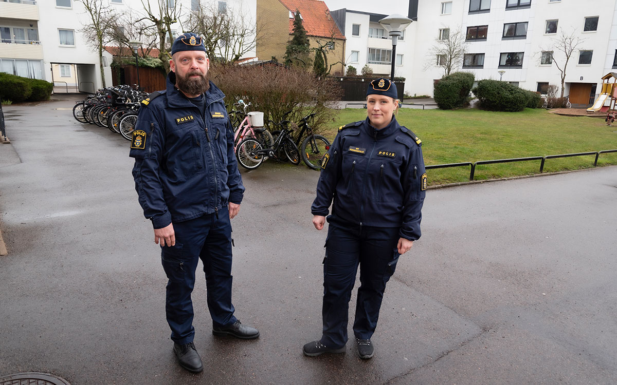 Två poliser, en man och en kvinna, i uniform och båtmössa som står på en innergård framför vita flerfamiljshus och har broderade skyltar på sina jackor, "Jörgen kommunpolis" och "My kommunpolis".