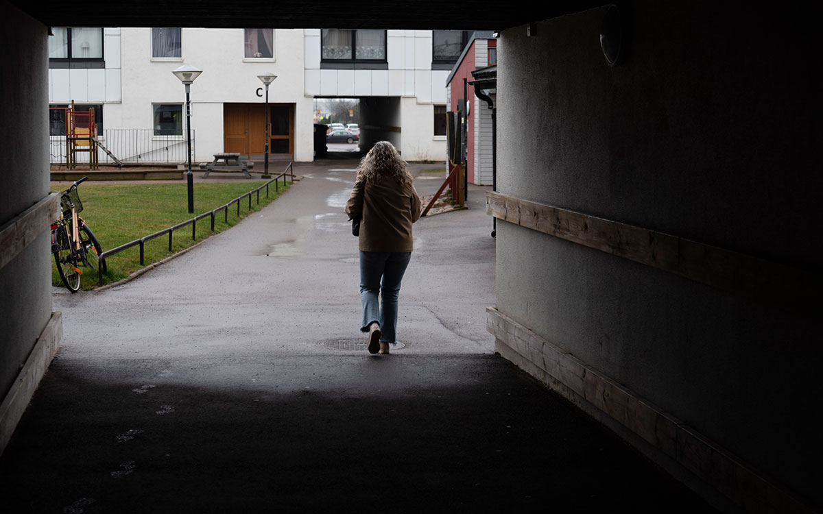 En kvinna med långt, grått hår som går igenom en öppning in på en innergård till en länga av vita flervåningshus.