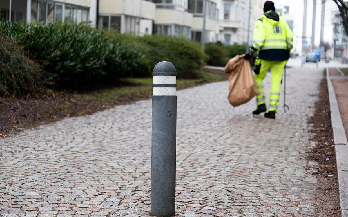 En stolpe i en stensatt gångbana och i bakgrunden en person i gula varselkläder och Landskrona kommuns stadsvapen på ryggen som plockar skräp med en skräpplockare och lägger i en brun säck.