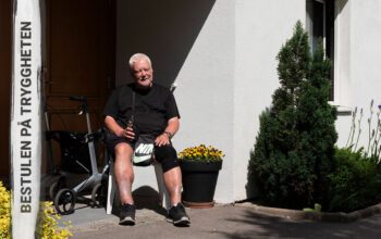 En äldre man med kortklippt vitt hår och svart t-tröja som sitter och dricker kaffe ur en mugg utanför en port och i bakgrunden syns en rullator och text på bilden "Bestulen på tryggheten".