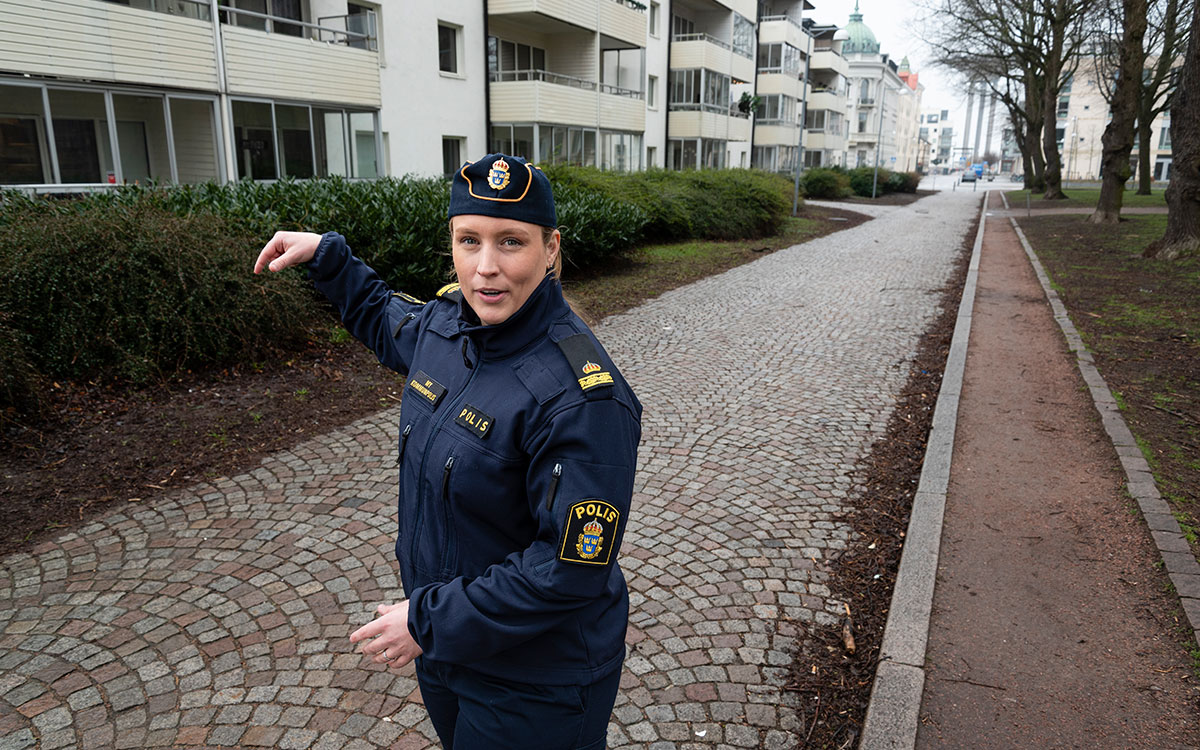 En ung kvinna i polisuniform med båtmössa och namnskylt med texten "My kommunpolis" och i bakgrunden skymtar ett vitt hyreshus och en stensatt gångväg och ett vattentorn.