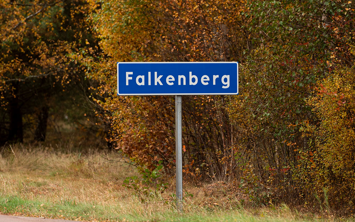 En blå och vit trafikskylt med texten "Falkenberg" som står framför höstgula björkar och i förgrunden en asfalterad landsväg.