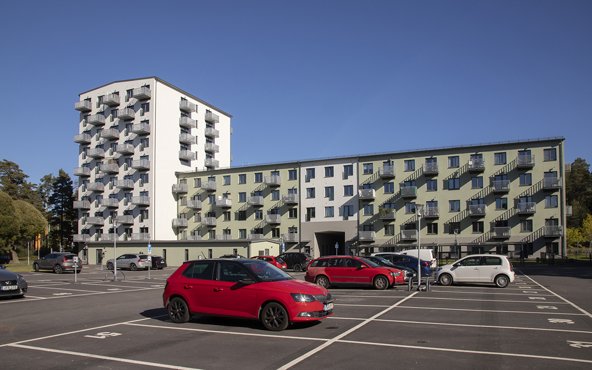 Fasadfoto på ett vitt höghus med många balkonger i grå metall och intill det ett ett ljust olivgrönt avlångt femvåningshus med en vitsektion i mitten. I förgrunden en röd bil på en parkering där de flesta parkeringsplatser är lediga. Joel Persson bor i en nybyggd lägenhet och har haft 14 grader hemma. Nu vill hyresvärden installera ett element som är så tjockt att hans kombinerade soffa och säng inte får plats i lägenheten.