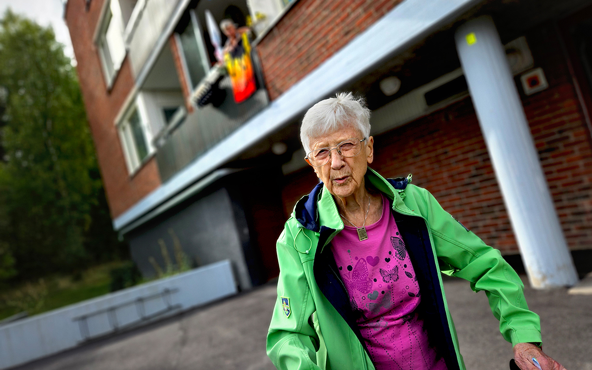 En kvinna som håller i handtagen på en rollator. I bakgrunden ett flerfamiljshus i tegel.