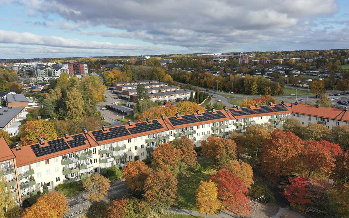 Foto ovanifrån på en lång huslänga i tre våningar med ljus fasad, gröna balkonger, tegelfärgat tak med solpaneler på. Rint omkring andra hus och träd i höstfärger. Husen hos Mimer är några av dem bolaget har med solceller. Många hyresvärdar har solceller, men än så länge är det ganska ovanligt att solelen går till hyresgästernas hushållsel trots att det kan sänka elräkningen både för hyresvärdarna och hyresgästerna.