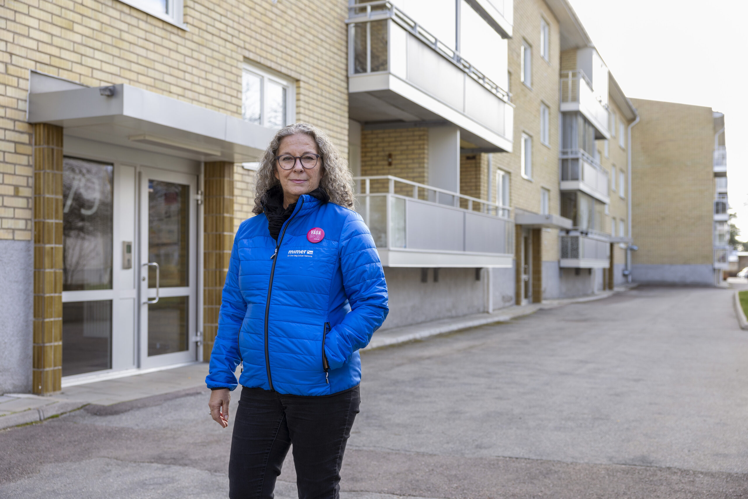 En kvinna i blå täckjacka och svarta byxor, grått lockigt hår ner över axlarna och glasögon. Hon står med vänstra handen i fickan framför en port till ett flerfamiljshus i ljusgult tegel.