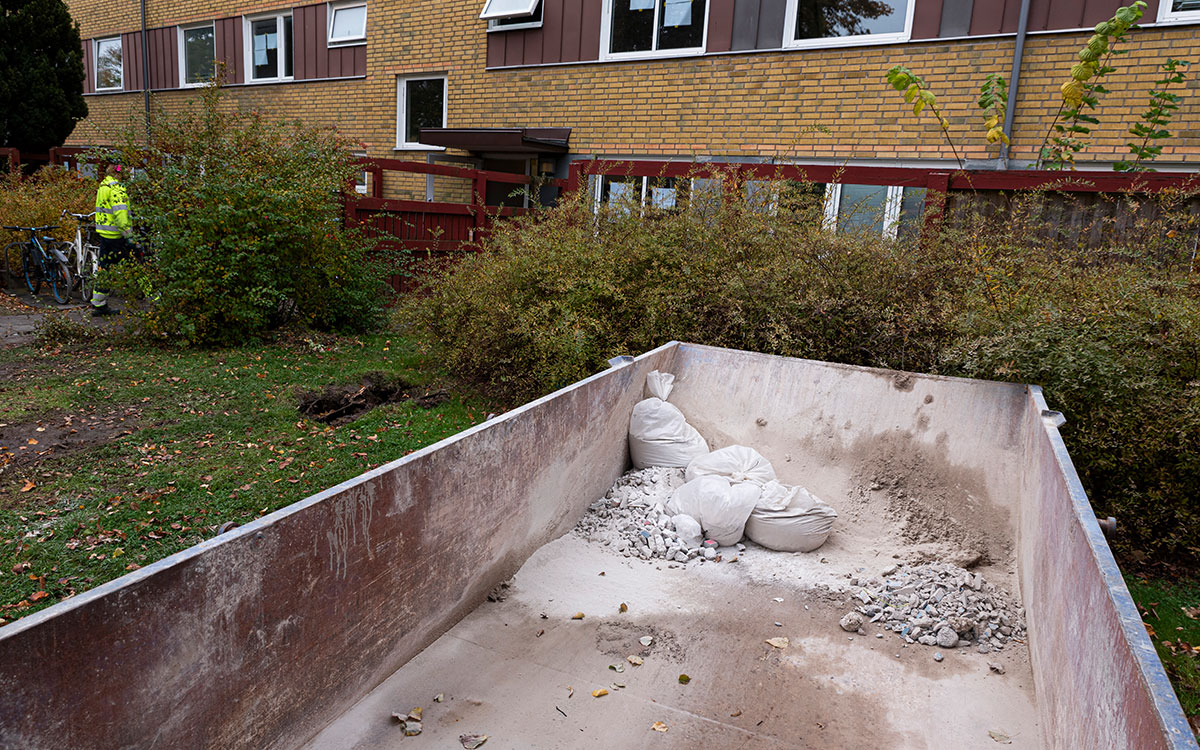 En container med säckar samt rester av betong, puts och cement som står framför ett flervåningshus i gult tegel och i bakgrunden en byggarbetare i gula varselkläder.