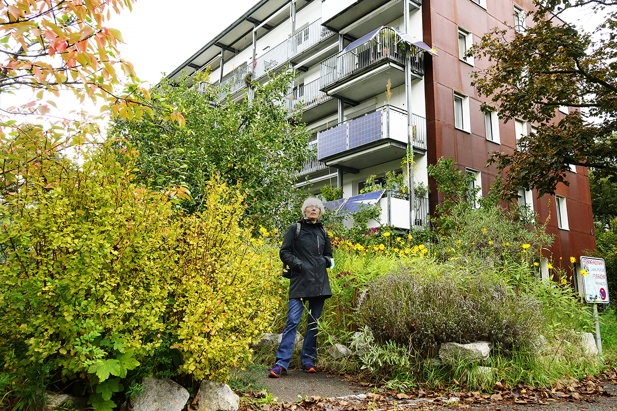 En kvinna som står framför ett hus och grönska växter och blommor.