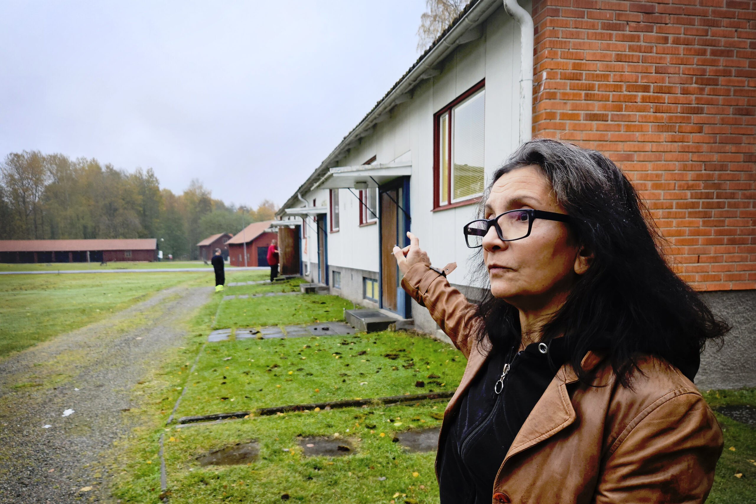 En kvinna med svart hår och glasögon pekar med armen mot ett hus.