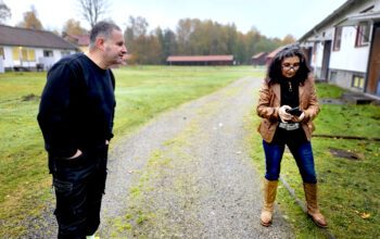 En man i svarta arbetskläder står på ena sidan av en väg. På andra sidan en kvinna med brun skinnjacka och bruna stövlar.