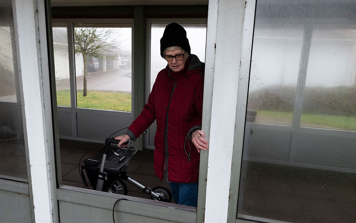 En äldre kvinna med rullator som sticker ut handen genom en fönsteröppning som saknar glasruta.