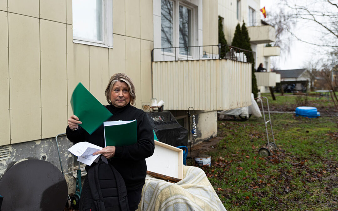 En kvinna med en jacka med texten "Hyresgästföreningen" som står framför ett gult hyreshus och i händerna håller flera papper och plastmappar och i bakgrunden skymtar en skånsk flagga.