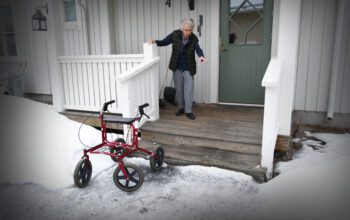 En bild på en äldre dam som håller sig i en stolpe med ena handen ovanför en trätrapp. Vänstra armen är utsträckt för att hålla balansen medan hon koncentrerat tittar ner på trappstegen. Nedanför trätrappan, som bara är några steg, står en rollator och väntar. Bilden är tagen utomhus.