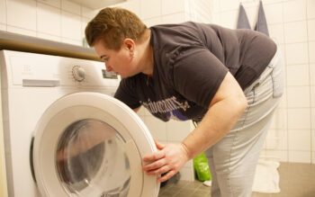 En person med kort hår brunrött hår, ring i örat, mörk t-shirt och ljusa jeans syns från sidan när hon böjer sig fram för att lägga in tvätt i tvättmaskinen. Uppsalahem återbrukar reservdelar, vilket gör att det kan hjälpa till snabbare vid felanmälningar. Nu ska de börja återbruka mer systematiskt och i större skala.