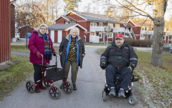 Tre personer står på en gångväg i ett bostadsområde med röda trähus i två våningar i bakgrunden. En äldre kvinna i grå page och vinröd kappa, randig halsduk och med rollator, en äldre kvinna i vit page, mörkbrun jacka, blå tröja ovanpå en mönstrad tröja, och en man i röd keps och svart jacka, i elrullstol. Hyresgäster hos AB-hem i Herräng har tröttnat på problemen med hyresvärden.