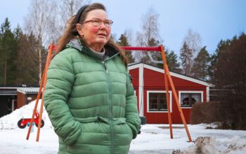 Kvinna i grön täckjacka som står med händerna i byxfickan. Hon tittar ut från bilden. I bakgrunden en liten lekpark. Då var hon medlem i områdets lokala hyresgästföreningen. I dag är den nedlagd.