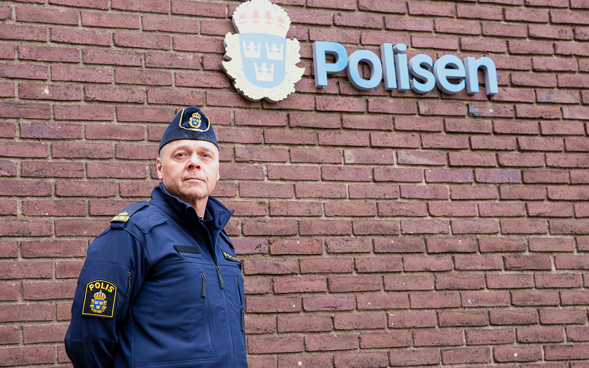 En polisman i uniform framför en röd tegelvägg. På väggen syns polisens emblem i blått och gult med de tre kronorna. Bredvid står det "Polisen".