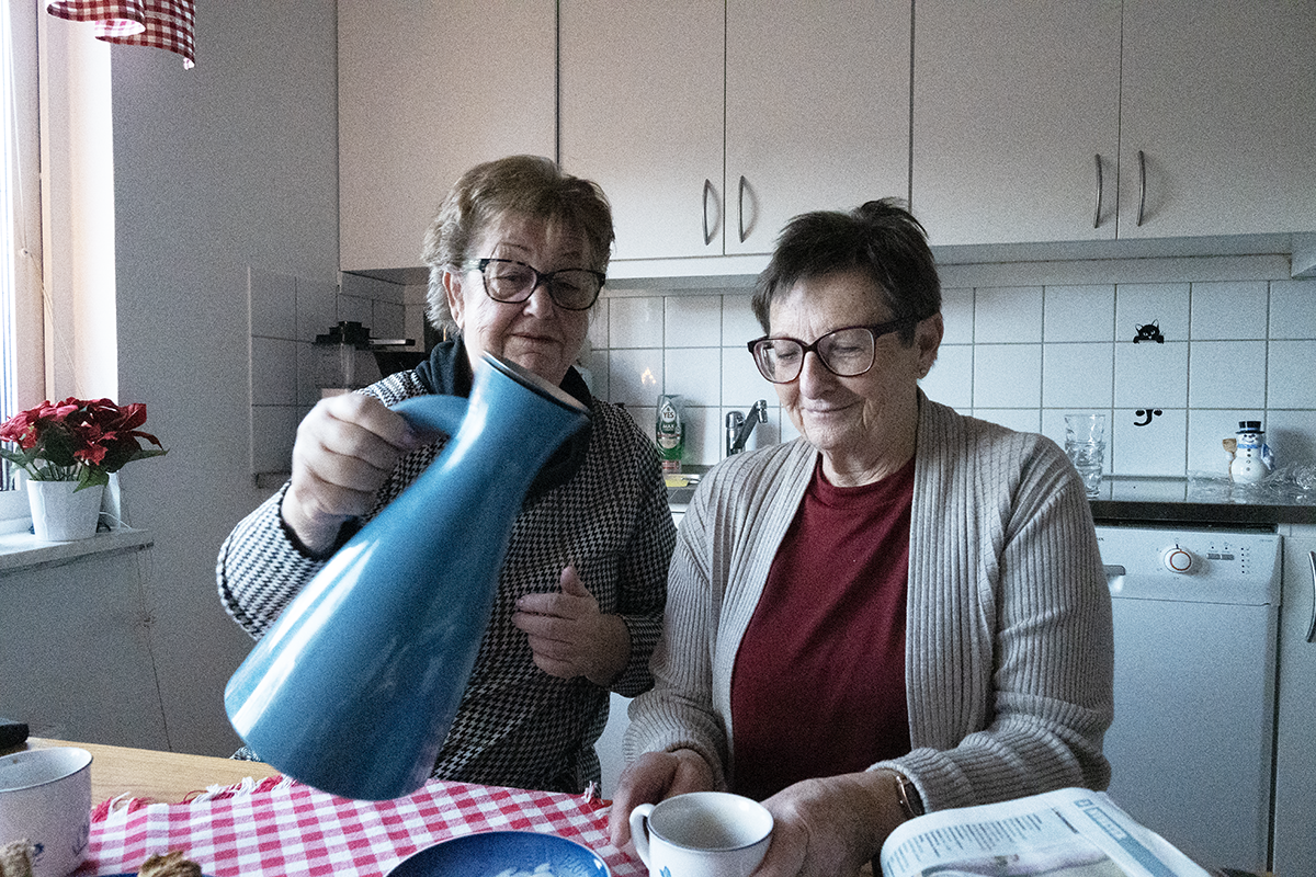 Två kvinnor i 70-årsåldern. En av dem häller upp kaffe från en blå kaffetermos.